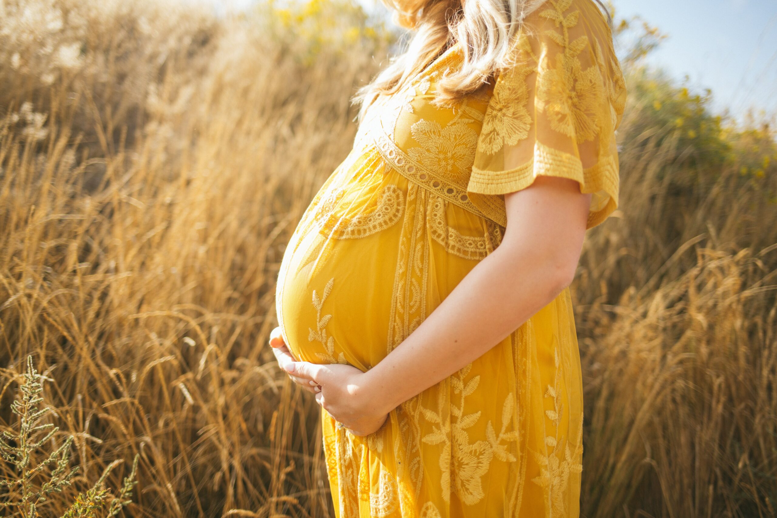 pregnant woman wearing yellow floral dress standing while touching her tummy and facing her right side near brown field during daytime